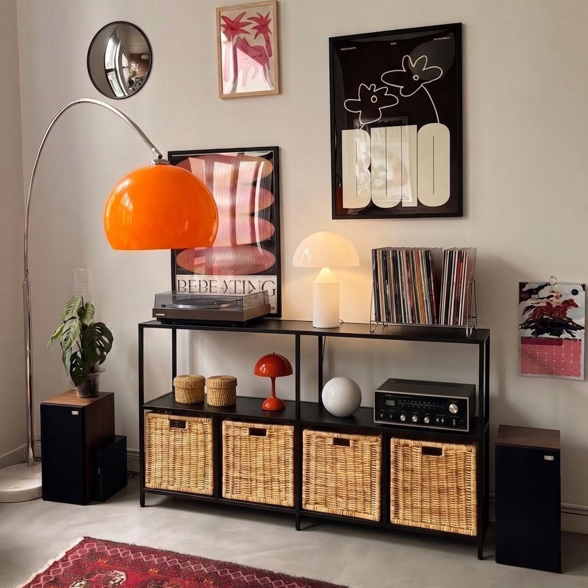 A styled interior scene with a black metal shelving unit holding woven storage baskets, records, a lamp, and decor, set against a light wall with framed art and a red patterned rug.